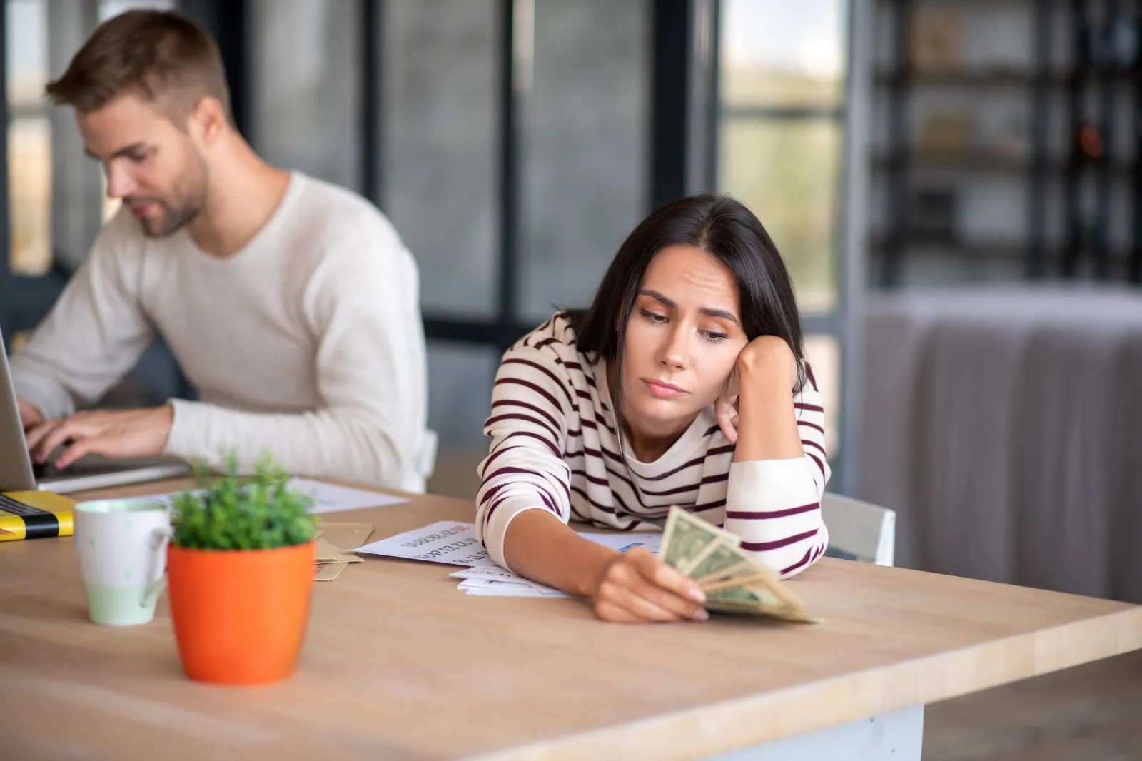 Woman looking concerned while counting cash at a table with paperwork and a man working on a laptop in the background, reflecting financial considerations related to family safety monitoring plans.