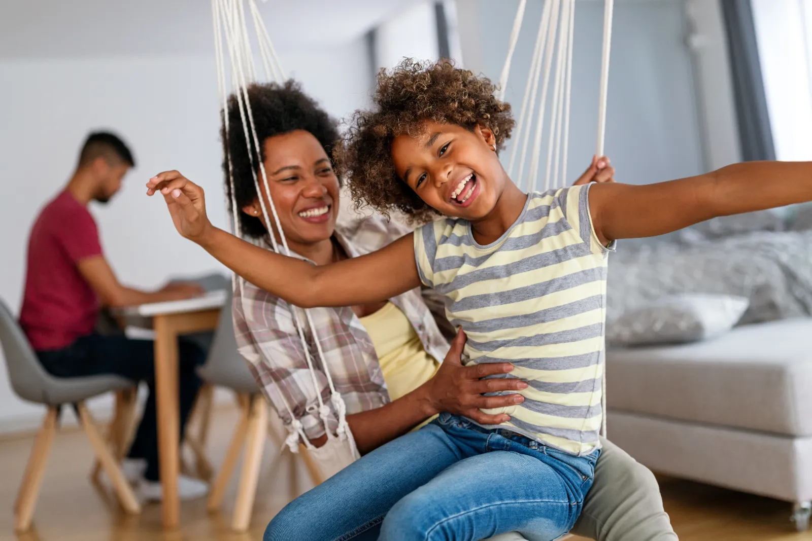 Mother and daughter enjoying playful moment in a swing, emphasizing family bonding and child safety in a cozy home environment, with a father working in the background.