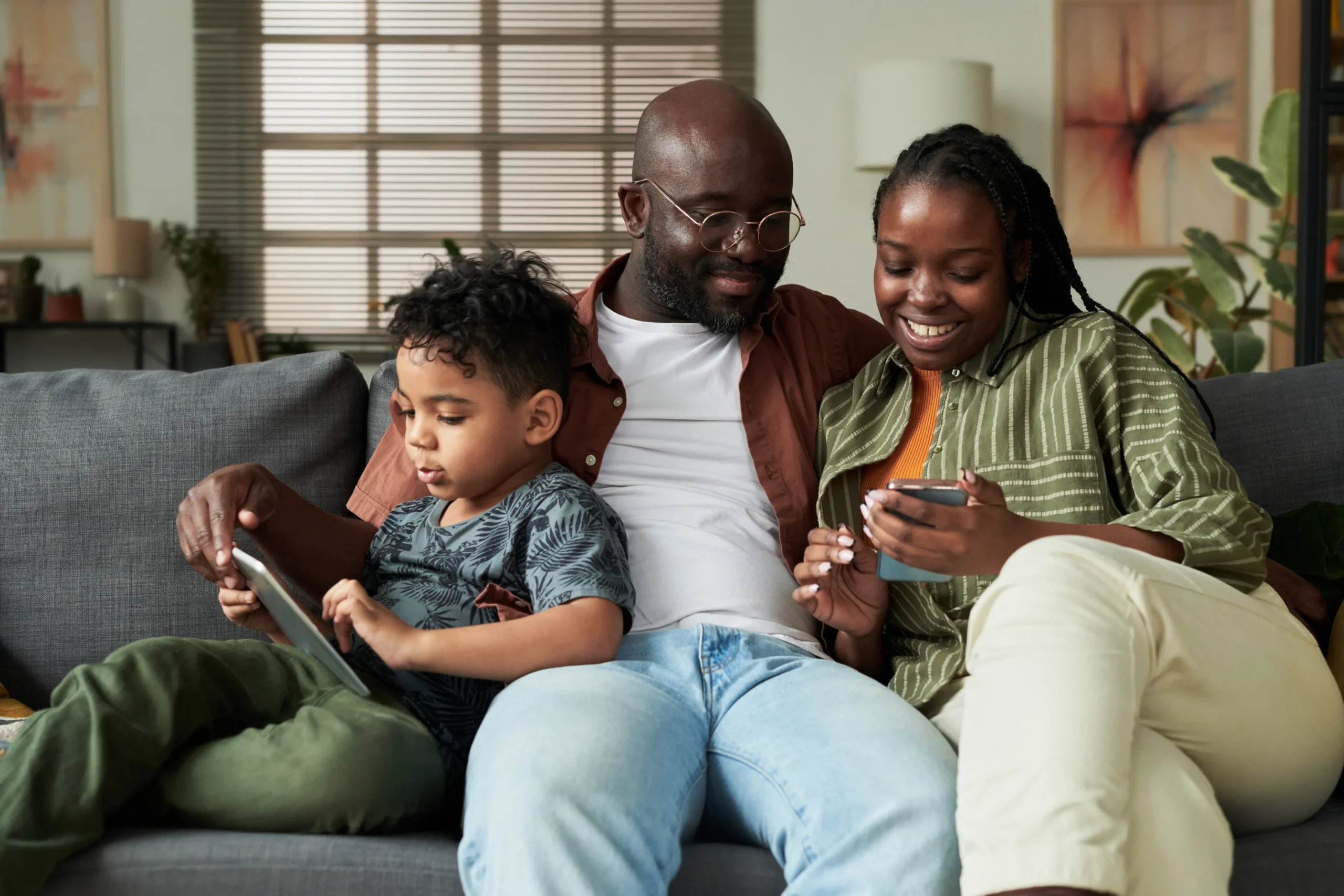 Family enjoying technology together on a couch, father using a smartphone, mother smiling while checking a phone, child focused on a tablet, emphasizing family connectivity and safety in digital environments.